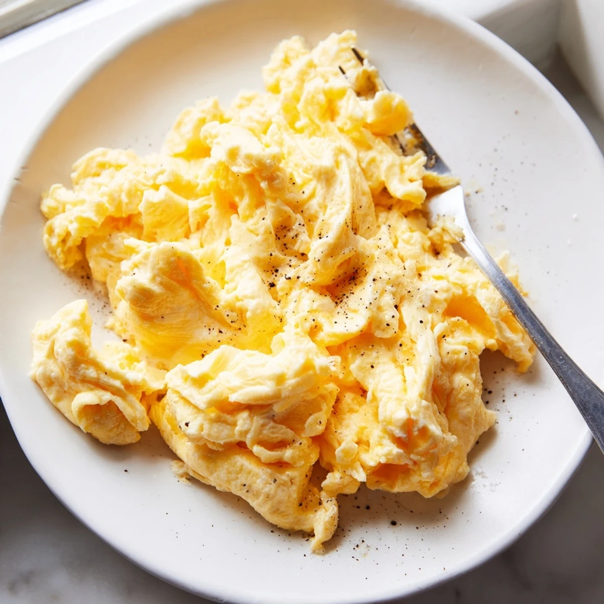 Creamy scrambled eggs piled on a white plate with buttery golden curds glistening under soft morning light